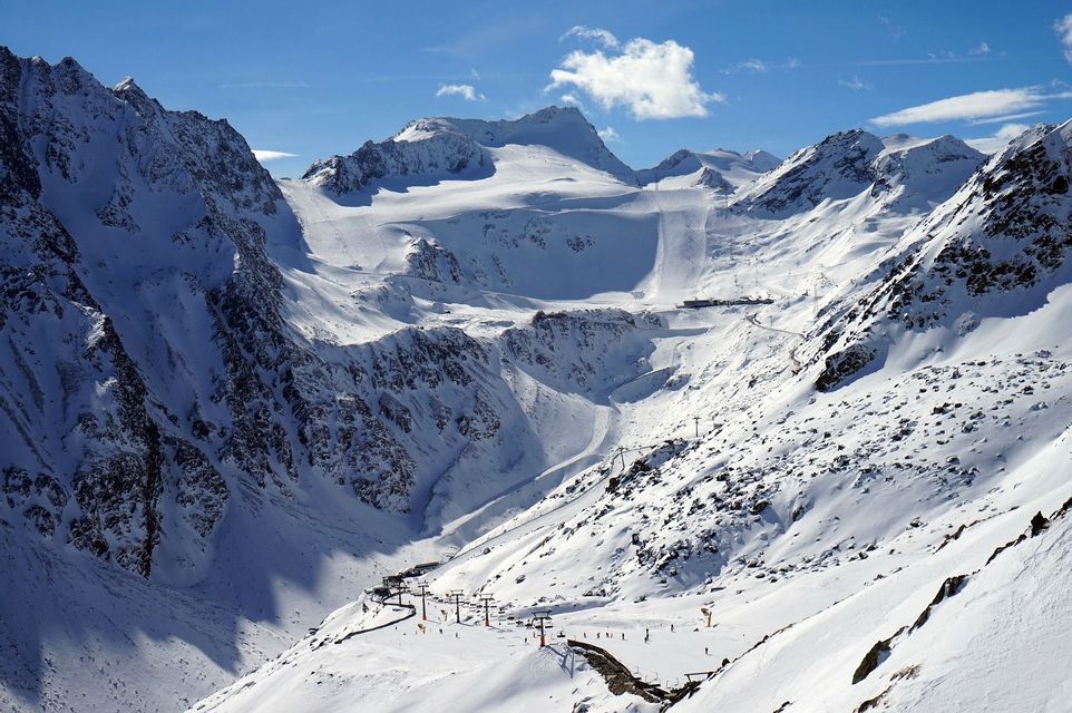 A vast, snow-covered mountain range with ski lifts and slopes visible under a bright blue sky with a few clouds.