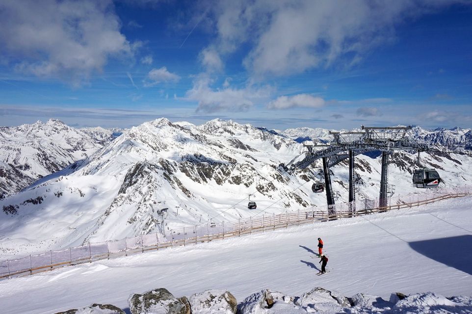 A wide view of a ski resort with two skiers on a slope, a gondola lift, and snow-capped mountains under a blue sky.
