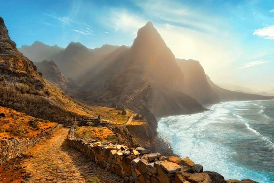 Ein gepflasterter Pfad folgt einer zerklüfteten, bergigen Küste, mit dem Meer auf der einen Seite und steilen Gipfeln im Hintergrund.
