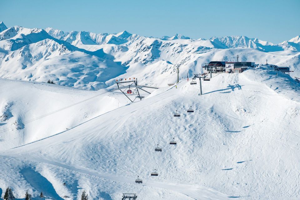 Sessellifte fahren einen sonnigen, schneebedeckten Berg zu einer Station hinauf, von der aus man einen Panoramablick auf Berggipfel hat.