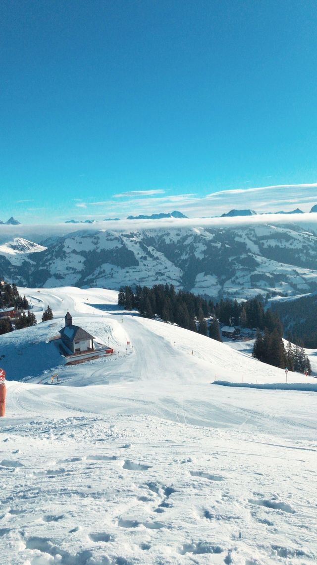 A small chapel sits next to a ski slope on a snow-capped mountain range under a bright blue sky.
