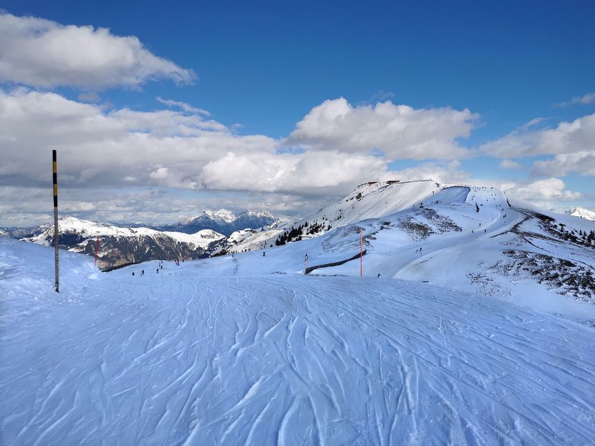 Ein schneebedeckter Berghang mit Skispuren im Vordergrund und entfernten Skifahrern unter blauem Himmel mit weißen Wolken.