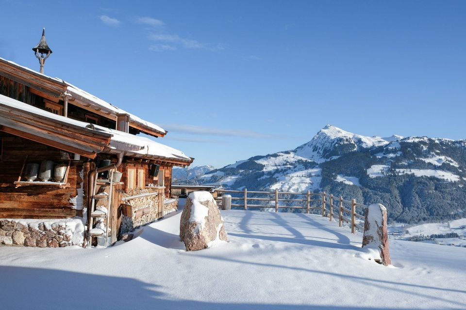 A wooden mountain cabin with a stone foundation is covered in snow, overlooking a snowy mountain range under a clear sky.