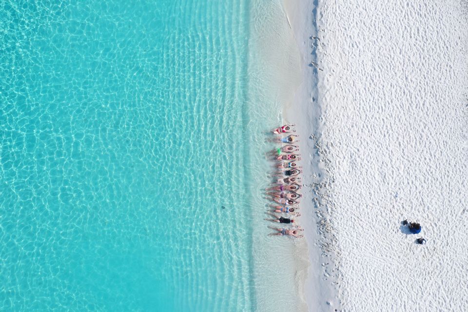 Vista aérea de un grupo de WeRoad tumbado en fila en una playa de arena blanca junto a aguas turquesas y cristalinas.