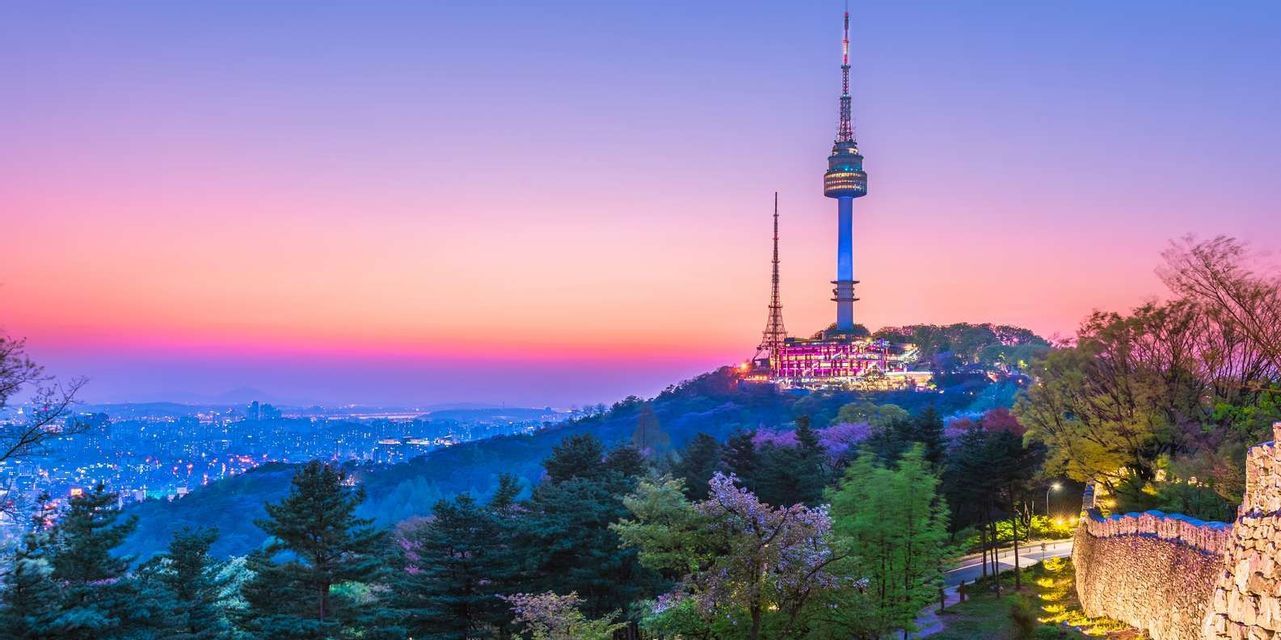 Una torre illuminata su una collina boscosa si affaccia su un vasto orizzonte urbano sotto un cielo al tramonto colorato.
