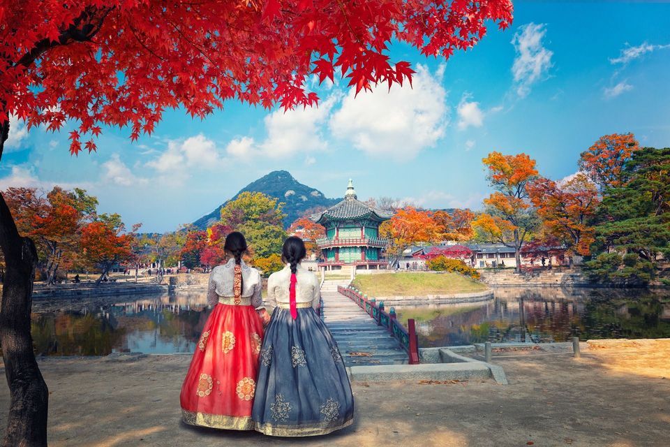 Due donne in abiti tradizionali coreani si trovano accanto a uno stagno, guardando una pagoda tra alberi autunnali colorati e montagne.