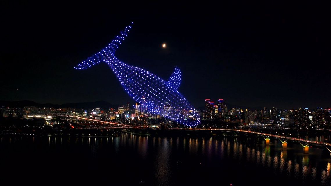 Uno spettacolo di luci con droni forma la sagoma di una grande balena blu nel cielo notturno su un paesaggio urbano illuminato con un fiume.