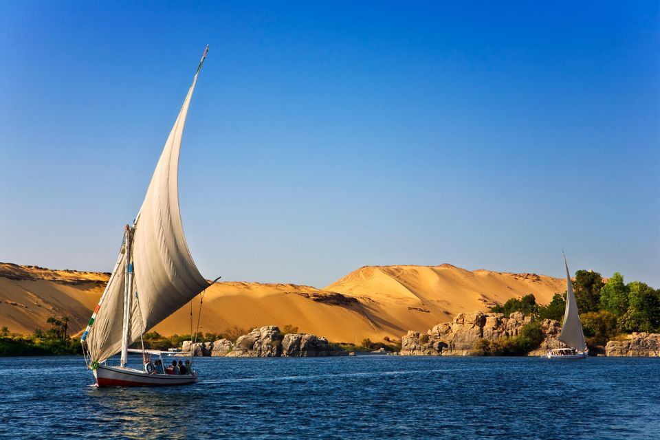Un viaggio di gruppo WeRoad naviga su una tradizionale feluca su un ampio fiume, oltre dune di sabbia dorata sotto un cielo azzurro chiaro.
