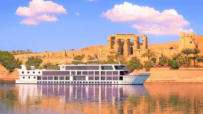 Una nave da crociera fluviale bianca galleggia sull'acqua davanti a rovine antiche su una riva sabbiosa con palme.