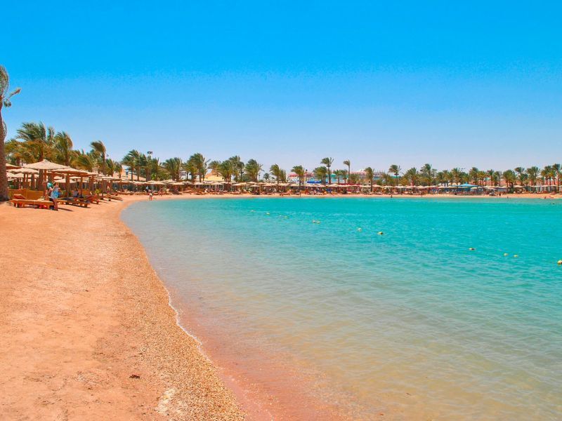 Una spiaggia di sabbia curva incontra acque calme e turchesi, con palme e ombrelloni di paglia che costeggiano la riva sotto un cielo limpido.