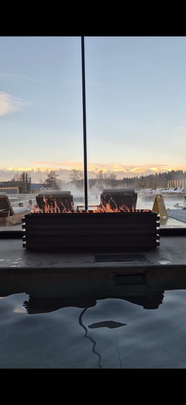 Una moderna hoguera arde junto a una humeante piscina termal exterior con bañistas, con un telón de fondo de montañas nevadas al anochecer.