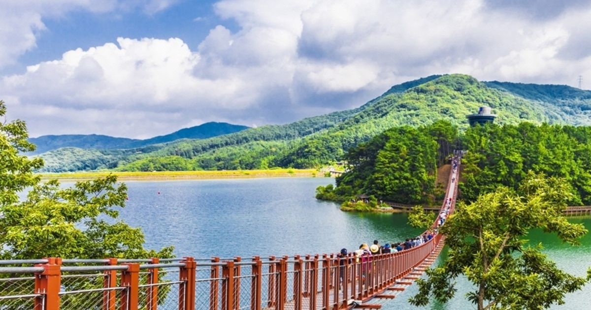 Persone che passeggiano su un lungo ponte sospeso sopra un lago, con montagne verdi e boscose sullo sfondo.
