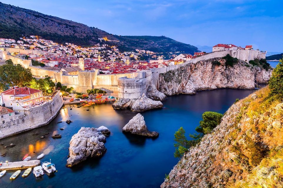 Una ciudad histórica amurallada en la costa se ilumina al anochecer, con vistas a una bahía tranquila con pequeños barcos.