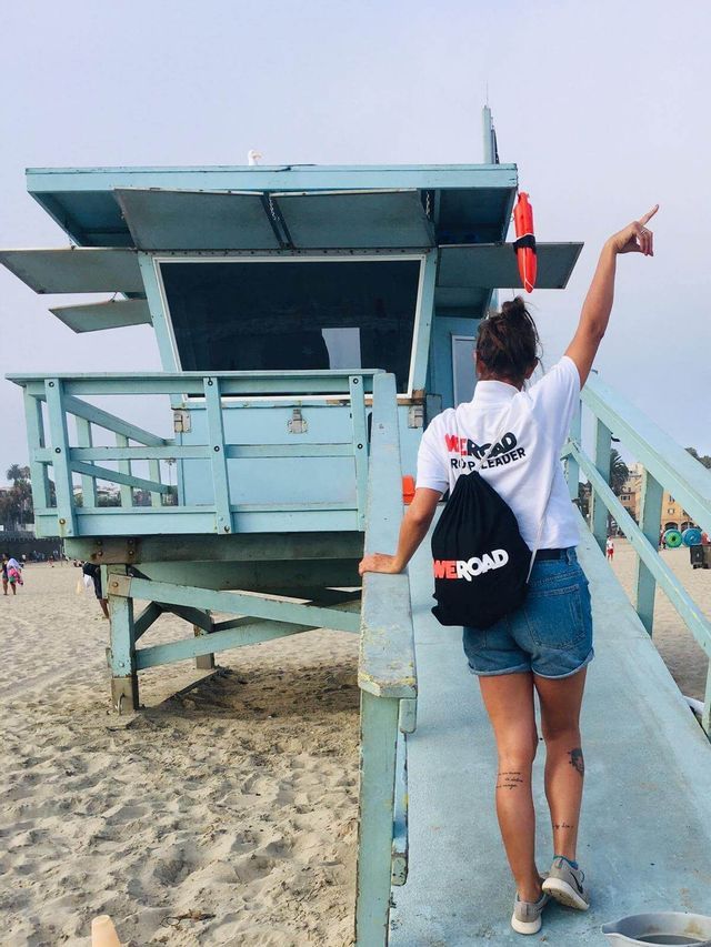A WeRoad group leader seen from behind stands on a ramp next to a lifeguard tower on a sandy beach, pointing upwards.