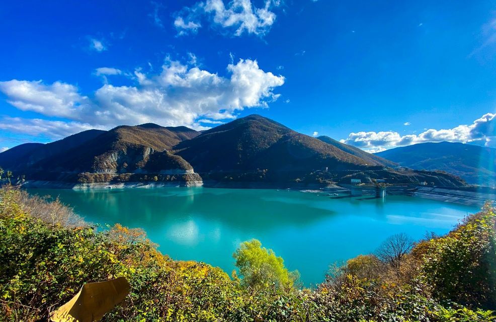 A vibrant turquoise lake sits between rolling mountains under a clear blue sky with some white clouds, seen from a high viewpoint.
