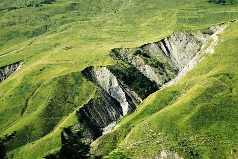 A deep ravine with exposed rock layers cuts through rolling hills covered in vibrant green grass.