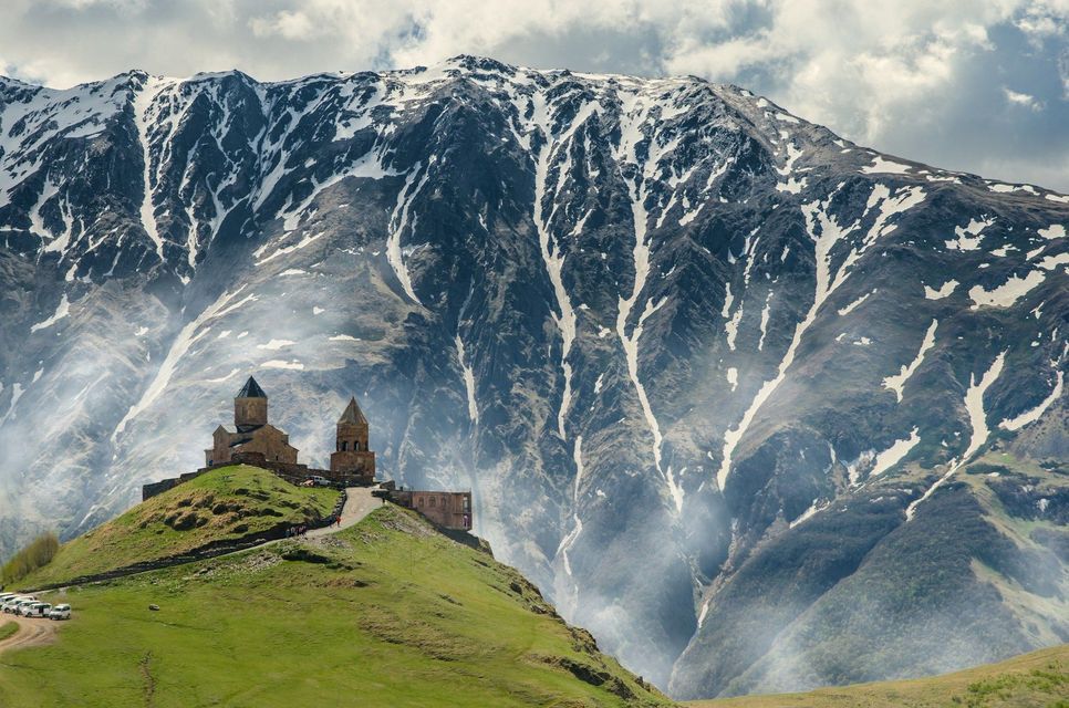 A stone church with two towers sits on a green hill with a large, snow-covered mountain in the background.