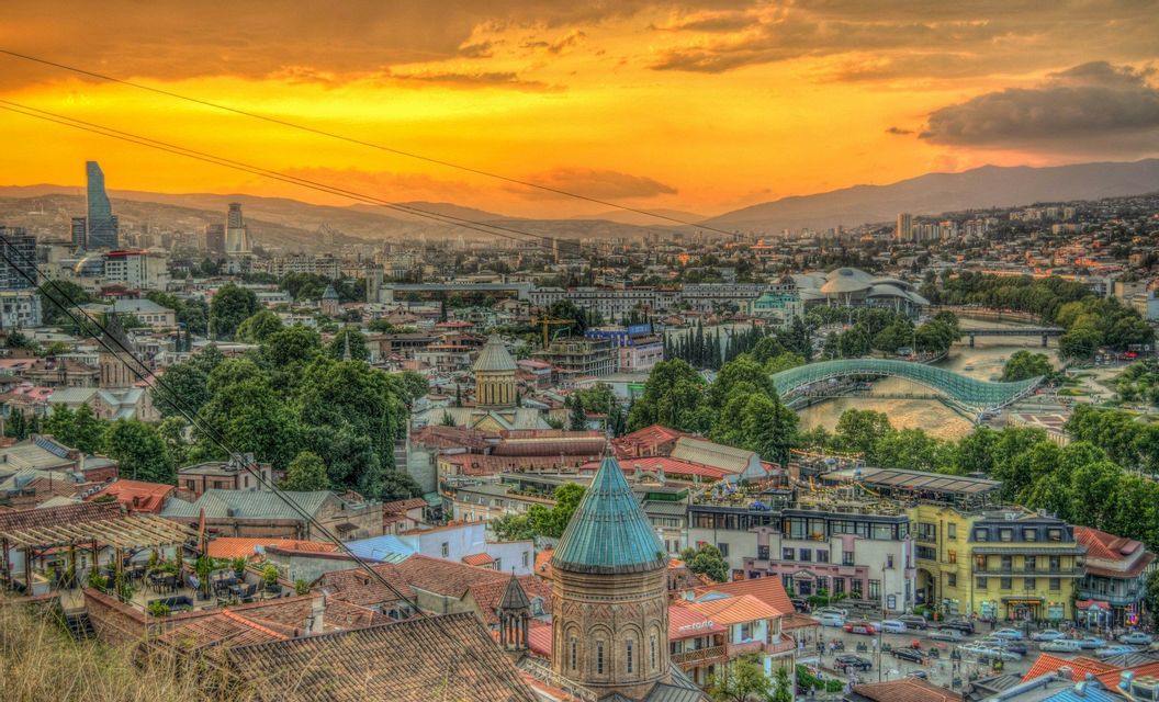 A panoramic view of a city with old churches, modern buildings, and a curved pedestrian bridge over a river, all under a golden sunset sky.