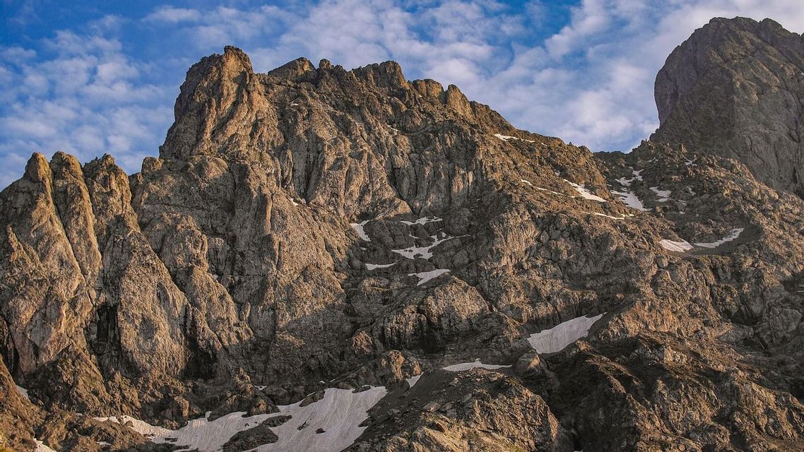 A rugged, rocky mountain peak with patches of snow on its slopes is illuminated by sunlight under a blue, cloudy sky.