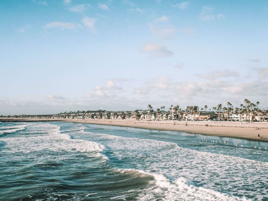 Olas del océano rompiendo en una amplia playa de arena, con una ciudad costera y palmeras visibles al fondo bajo un cielo azul.