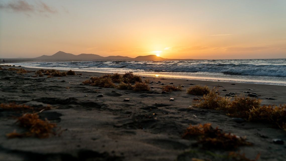Una vista dal basso di una spiaggia sabbiosa con alghe, mentre il sole tramonta sull'oceano e sulle montagne lontane.