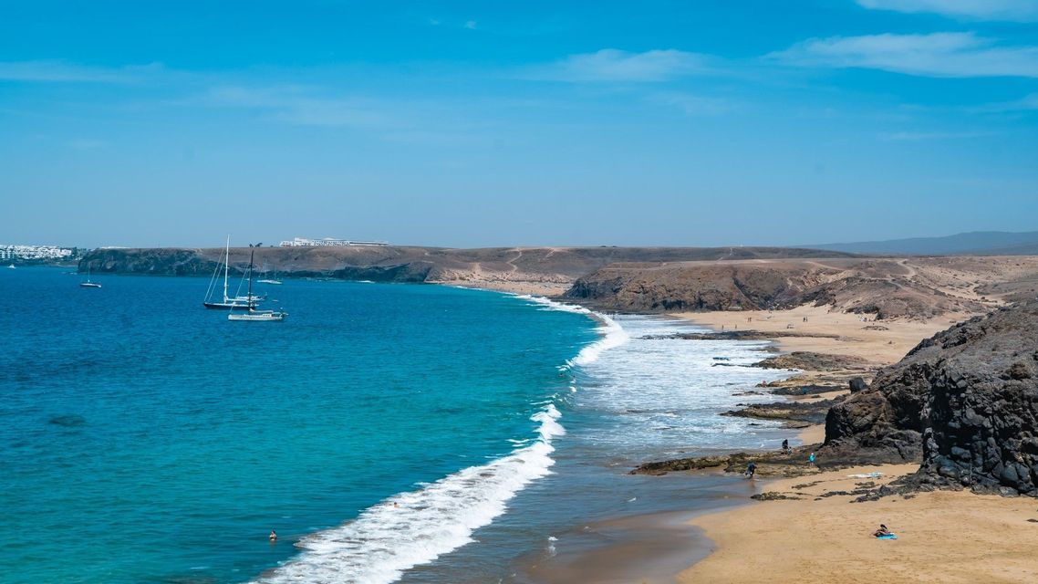 Una veduta aerea di una costa con acqua turchese che accarezza una riva sabbiosa e rocciosa, con diverse barche a vela ancorate nella baia.