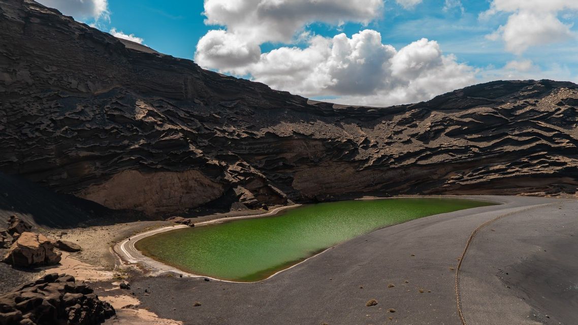 Un lago di un verde brillante incastonato alla base di un cratere vulcanico scuro e roccioso, sotto un cielo azzurro parzialmente nuvoloso.