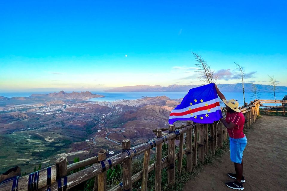 Eine Person mit Strohhut hält die Flagge Kap Verdes an einem Aussichtspunkt mit Blick auf eine Küstenstadt und Berge.