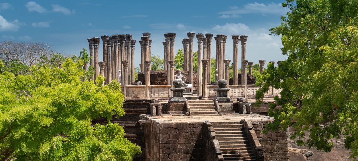 Las ruinas de un antiguo templo con pilares de piedra y estatuas están enmarcadas por frondosos árboles bajo un cielo azul.