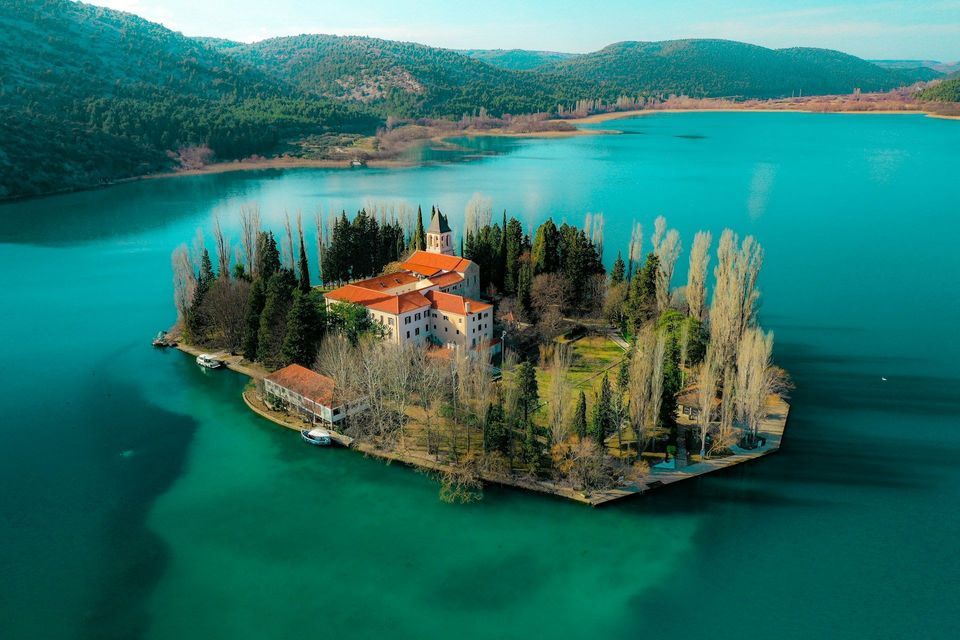 Luftaufnahme eines Klosters mit roten Dächern auf einer kleinen Insel in einem türkisfarbenen See, umgeben von bewaldeten Hügeln.