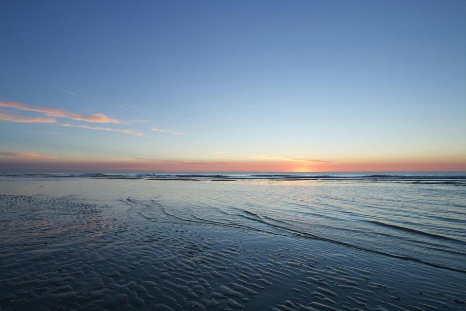 Die Sonne sinkt am Horizont über dem Ozean und wirft einen Schein auf den gewellten Sand eines feuchten Strandes bei Ebbe.