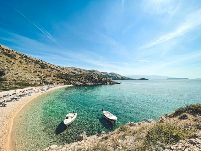 Eine Draufsicht auf eine sandige Bucht mit klarem, türkisfarbenem Wasser, zwei geankerten Booten und Personen am Strand.