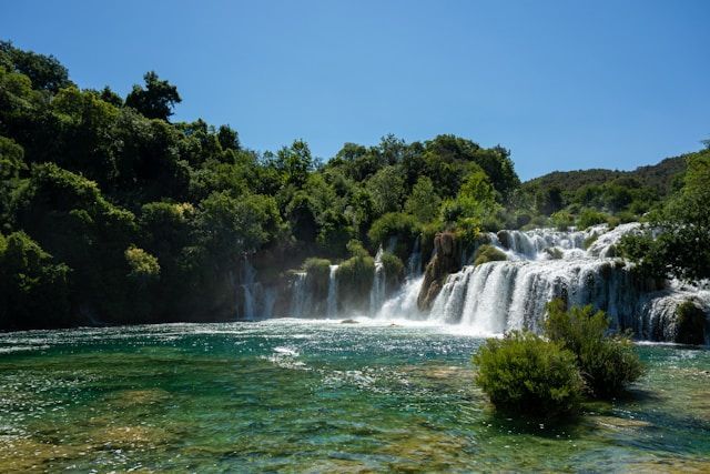 Ein mehrstufiger Wasserfall stürzt in einen klaren türkisfarbenen Fluss, umgeben von üppigem grünen Wald unter einem strahlend blauen Himmel.