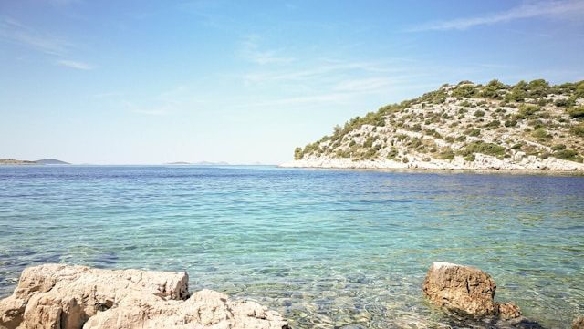 Klares türkisfarbenes Wasser in einer felsigen Bucht mit einem mit Buschwerk bewachsenen Hügel im Hintergrund unter einem strahlend blauen Himmel.
