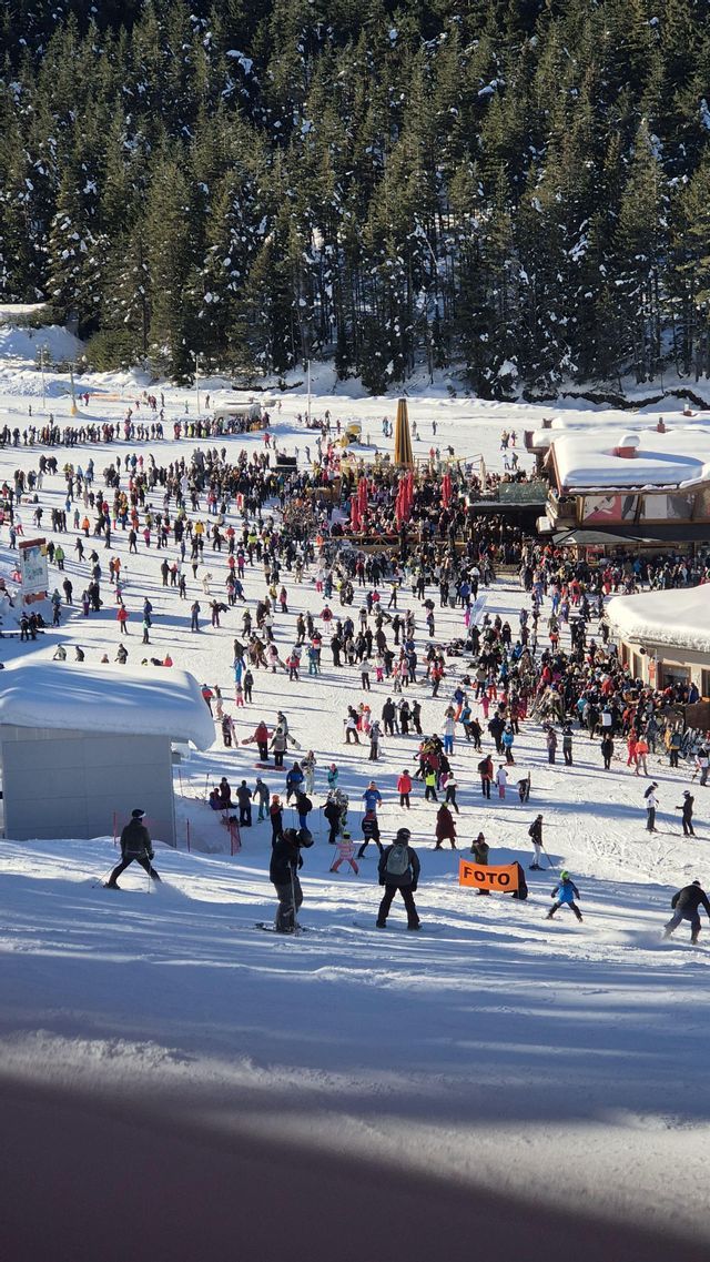 Panoramablick auf eine belebte Skipiste mit vielen Skifahrern und Snowboardern nahe einer Skihütte an einem sonnigen Tag.