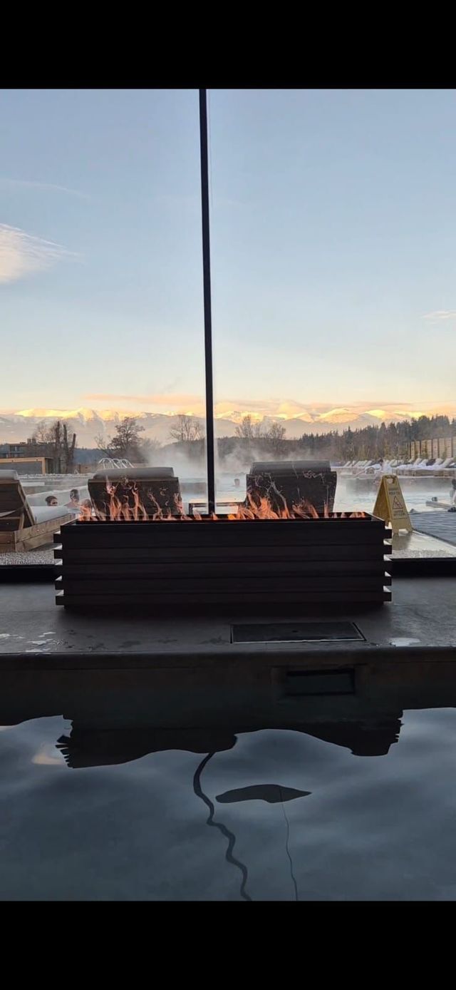 A fire pit burns beside a steaming outdoor thermal pool, with distant snow-capped mountains visible at dusk.