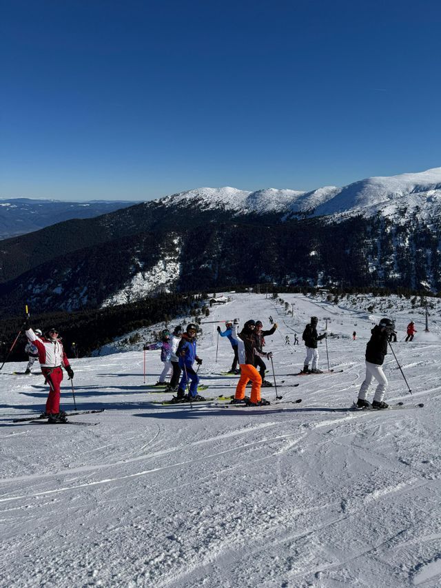 Un groupe WeRoad posant à ski sur une pente de montagne enneigée, sous un ciel bleu clair, avec des montagnes boisées en arrière-plan.