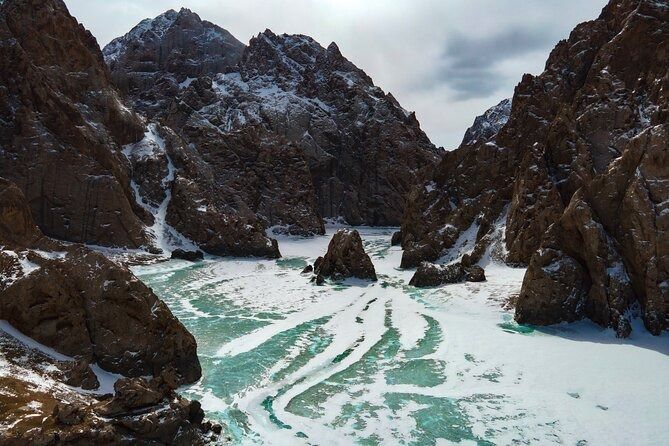 A frozen river with patches of turquoise ice winds through a steep, snow-dusted rocky canyon.