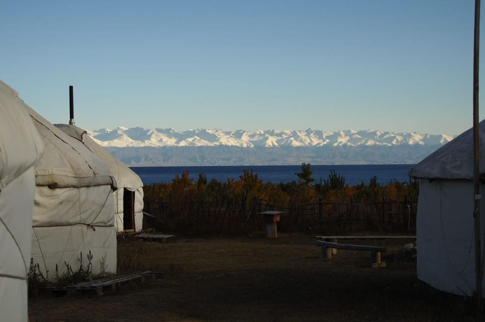 Deux yourtes au bord d&#39;un grand lac, avec une chaîne de montagnes enneigées visible au loin sous un ciel bleu clair.