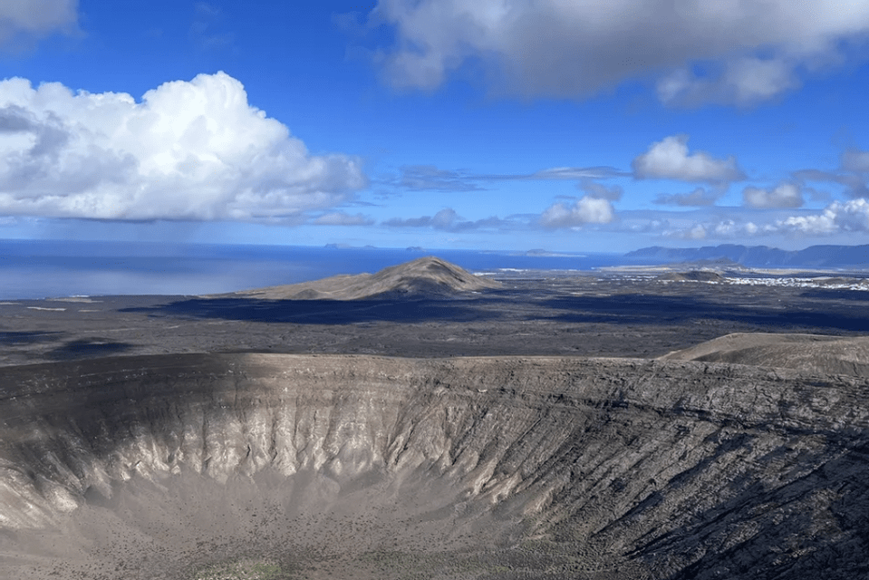 Una vista panorámica de un cráter volcánico en un vasto y oscuro paisaje, con el océano visible a lo lejos bajo un cielo azul y nublado.