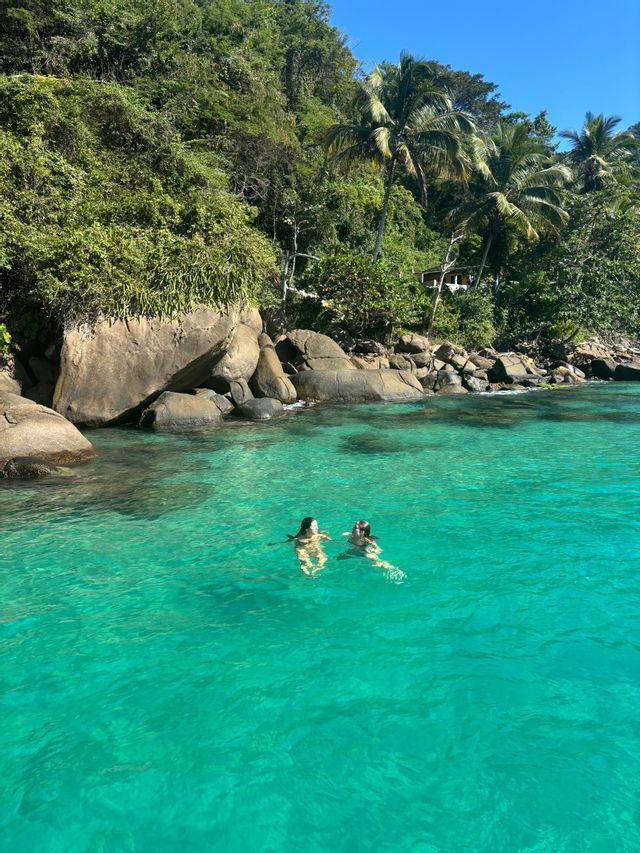 Zwei Personen einer WeRoad-Gruppenreise schwimmen im klaren, türkisfarbenen Wasser vor einer felsigen Küste mit üppigen grünen Bäumen.