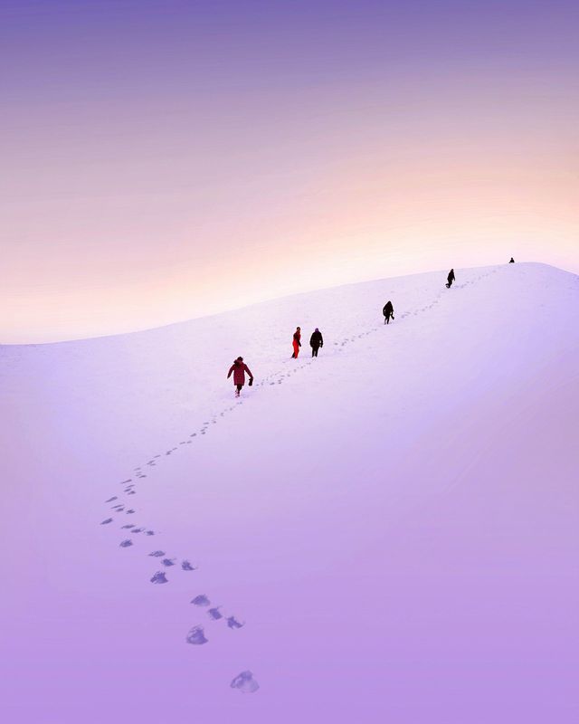 Un viaggio di gruppo WeRoad cammina in fila indiana su un vasto pendio innevato, lasciando impronte sotto un cielo viola e arancione.