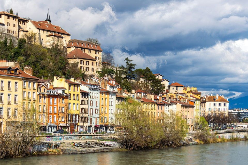 A row of colorful buildings stands on the bank of a river, at the foot of a green hill, under a partly cloudy sky.