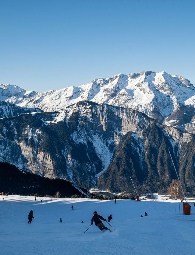Des skieurs descendent une pente enneigée par une journée ensoleillée, avec de grandes montagnes enneigées sous un ciel clair.