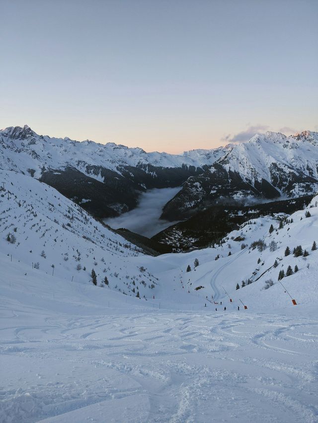 Un versant de montagne enneigé avec des pistes de ski surplombe une vallée enveloppée d&#39;une épaisse couche de nuages au coucher du soleil.
