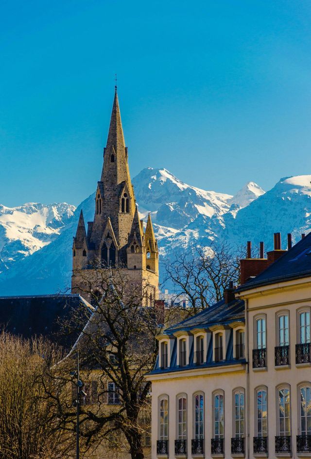 Une flèche d'église en pierre s'élève au-dessus des toits de la ville, avec un arrière-plan de montagnes enneigées sous un ciel bleu clair.