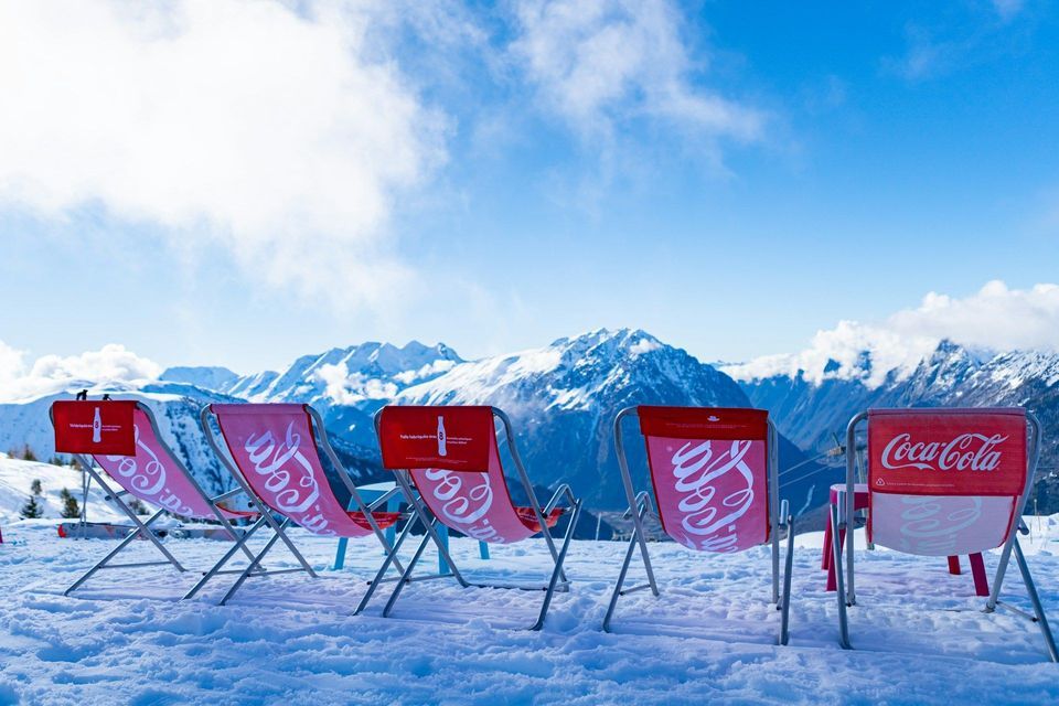 A row of red Coca-Cola branded deck chairs sit empty in the snow, facing a mountain range under a blue sky.
