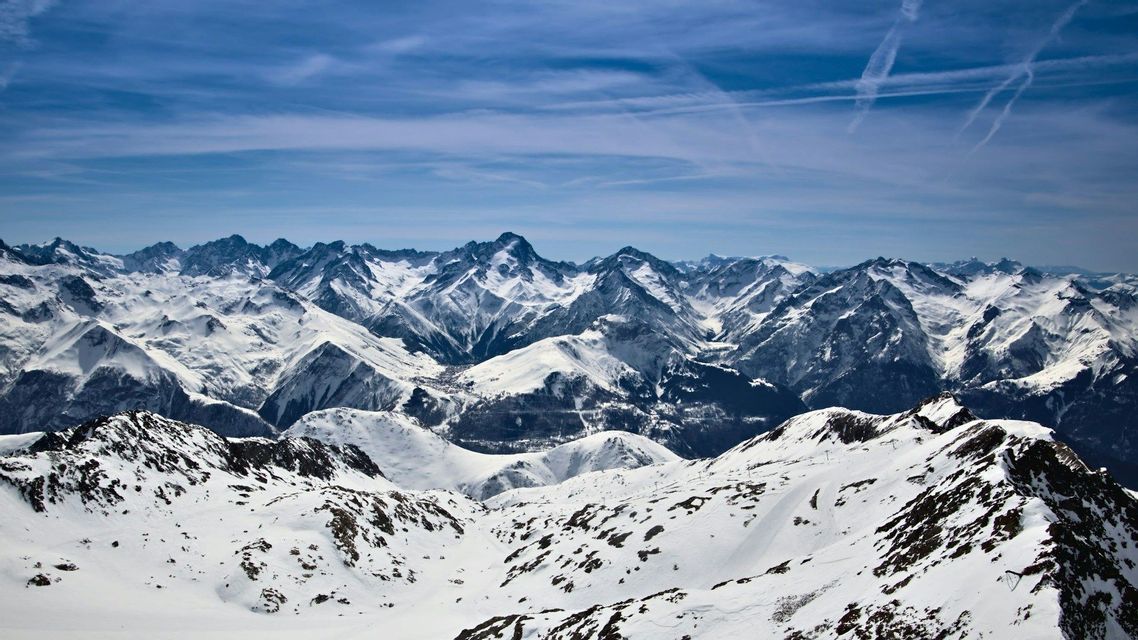 Une vue panoramique d&#39;une vaste chaîne de montagnes enneigées sous un ciel bleu avec des nuages filamenteux et des traînées de condensation.