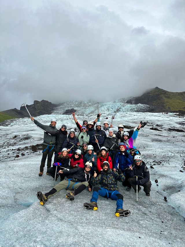 A WeRoad group trip poses for a photo on a glacier, equipped with helmets, crampons, and ice axes under a cloudy sky.