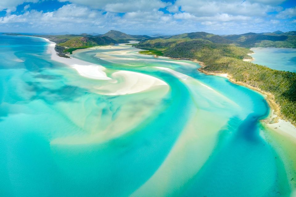 Una vista aerea di banchi di sabbia bianchi e vorticosi in acque turchesi vibranti, con isole verdi e collinari sullo sfondo sotto un cielo blu.